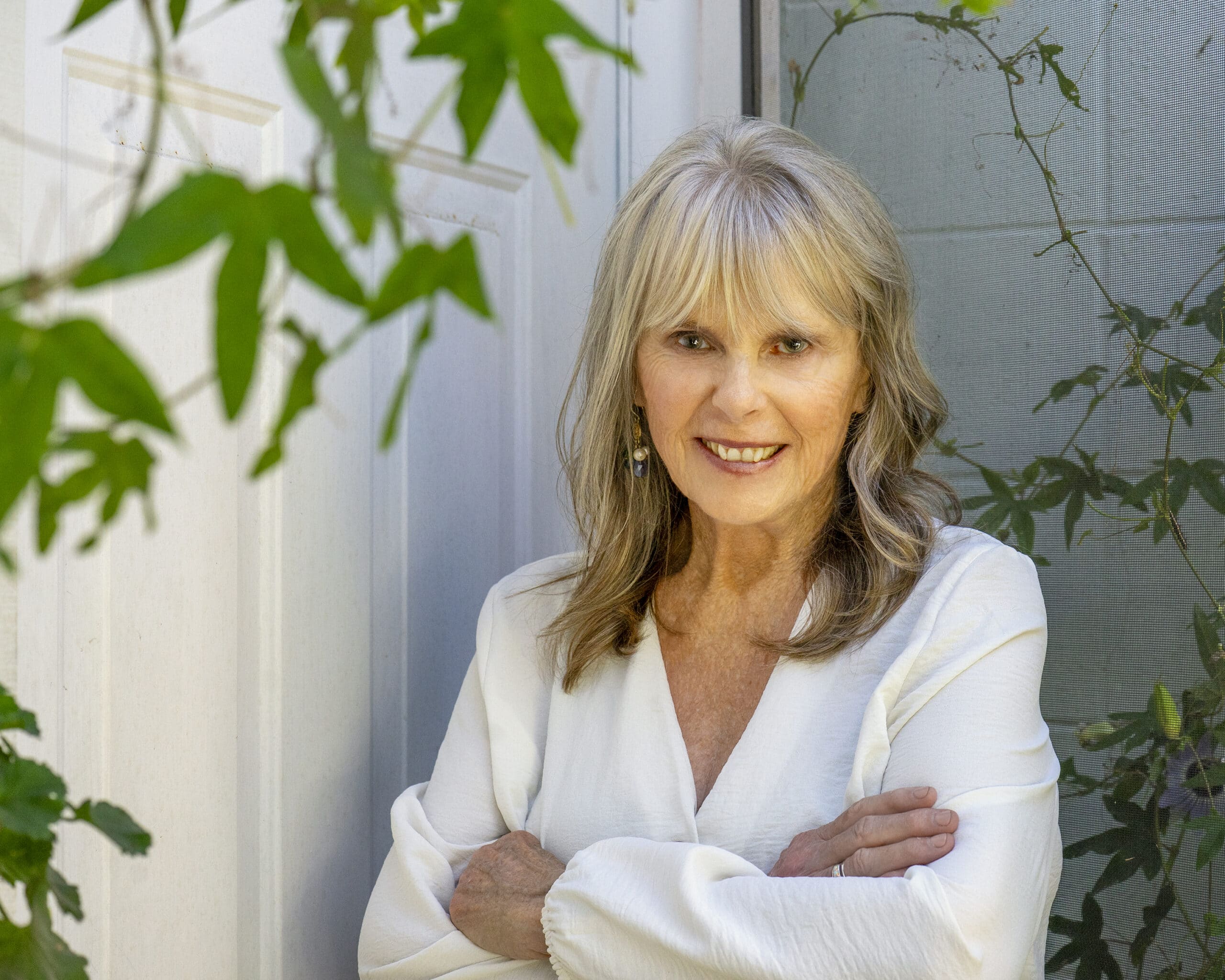 Dr. Susan Allison, Ph.D., in a white linen blouse with arms folded, among green vines