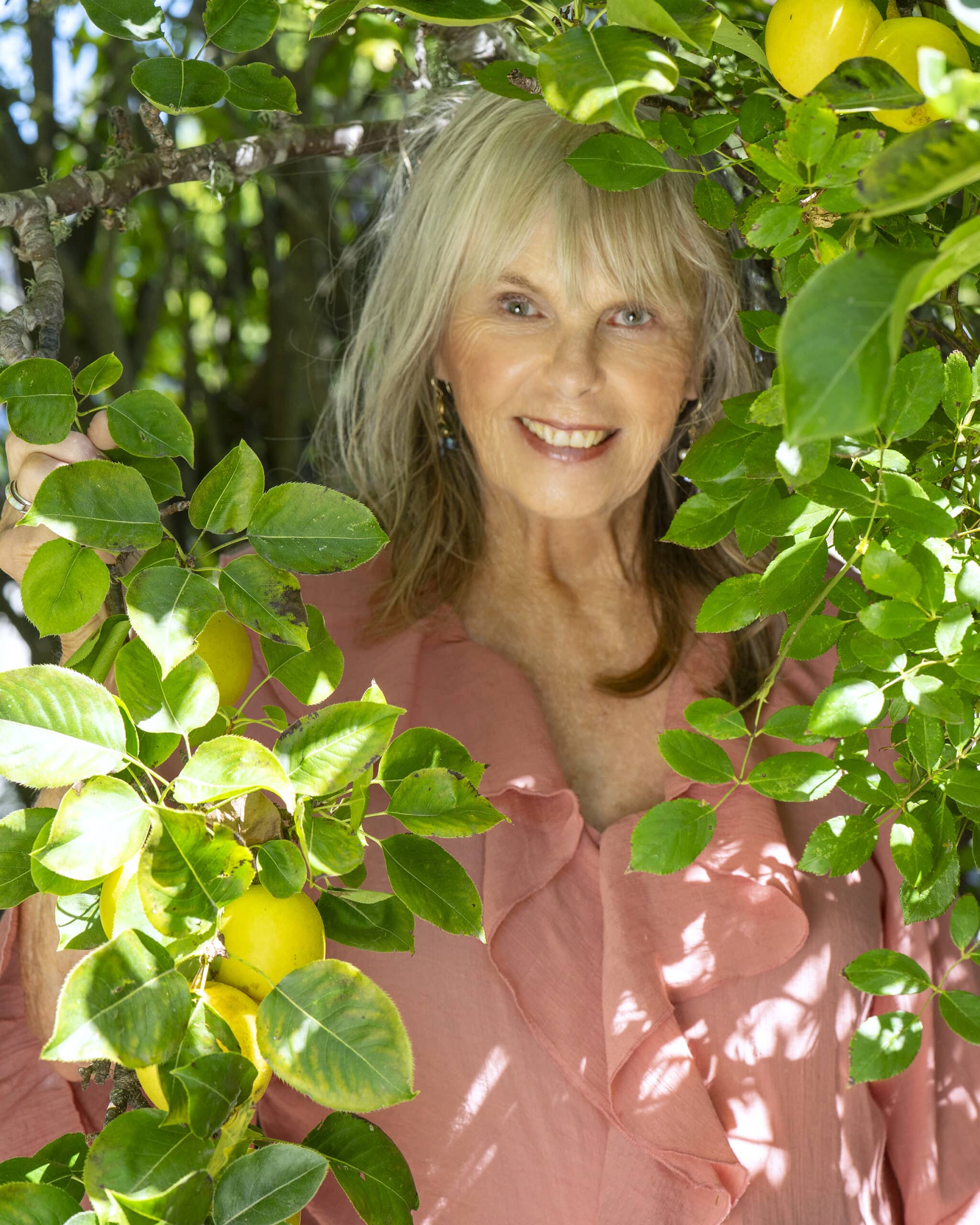 Dr. Susan Allison, Ph.D., in a pink ruffled blouse, smiling through green leaves and yellow fruit