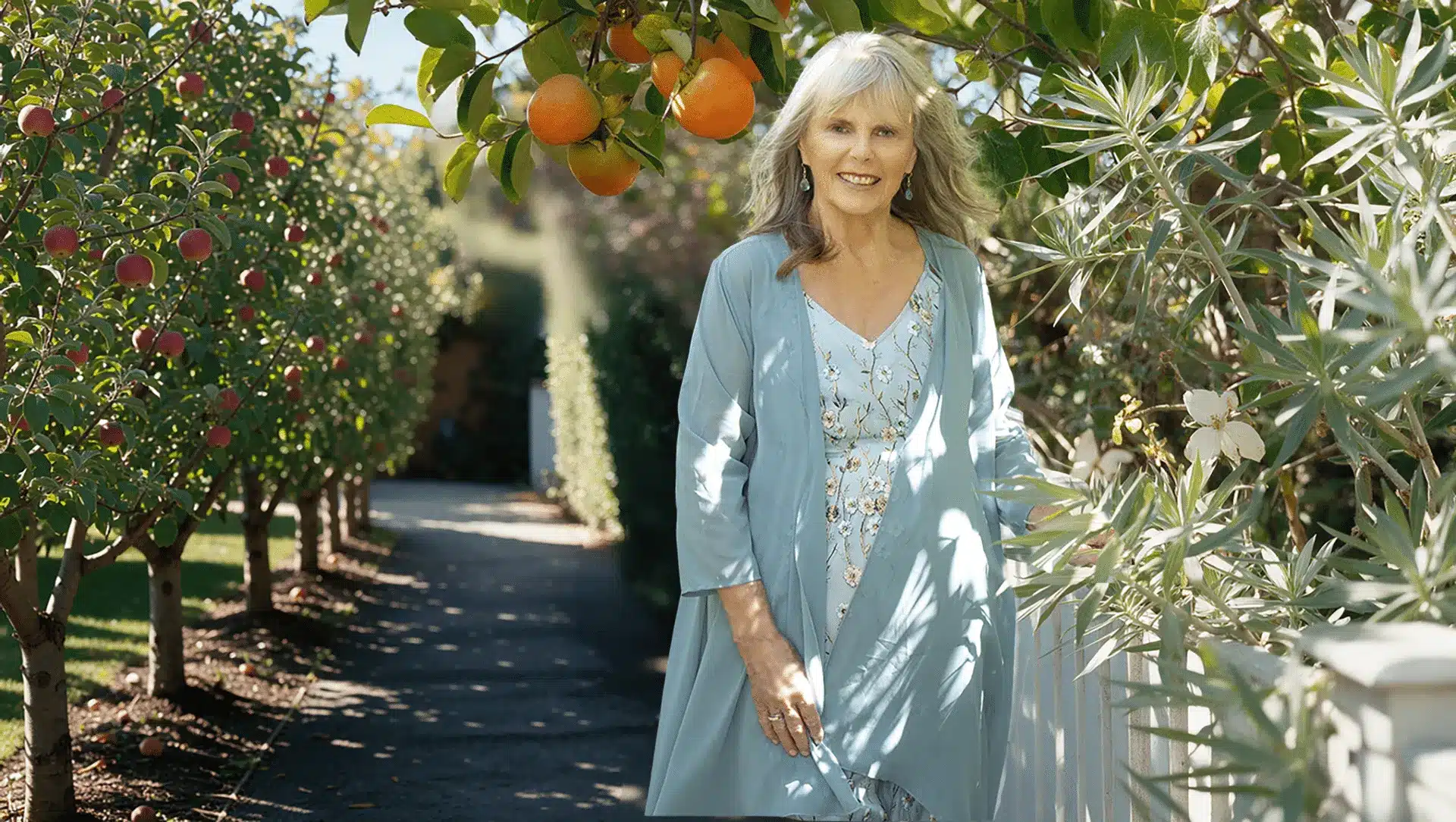 Dr. Susan Allison, Ph.D., smiling as she walks an orchard path lined with apple trees and a persimmon branch overhead