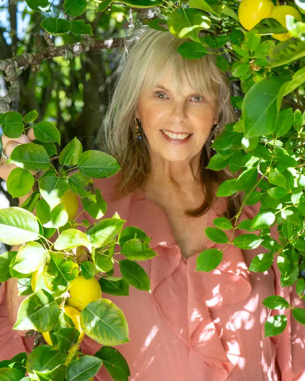 Dr. Susan Allison, Ph.D., in a pink ruffled blouse, smiling through green leaves and yellow fruit