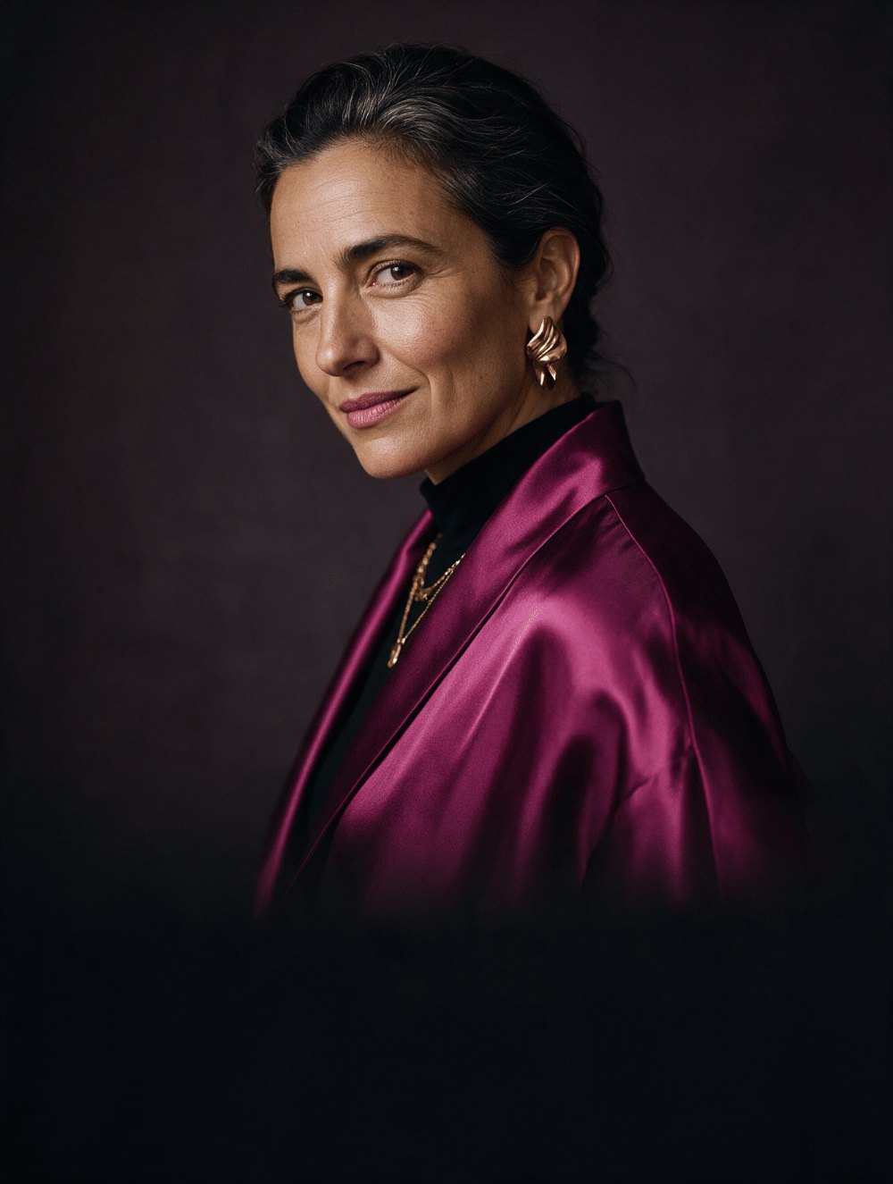 Portrait of a woman in a magenta satin jacket and gold earrings, looking over her shoulder at the camera